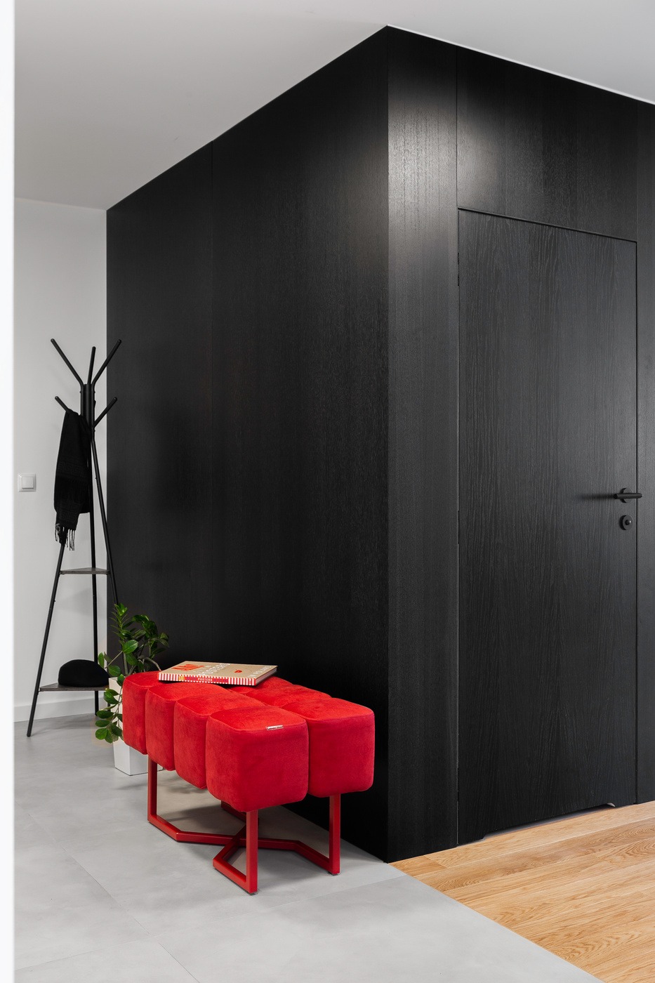 Nowoczesny przedpokój z czarną zabudową, ukrytymi drzwiami i czerwonym pufem na metalowym stelażu, uzupełniony wieszakiem na ubrania i rośliną doniczkową.