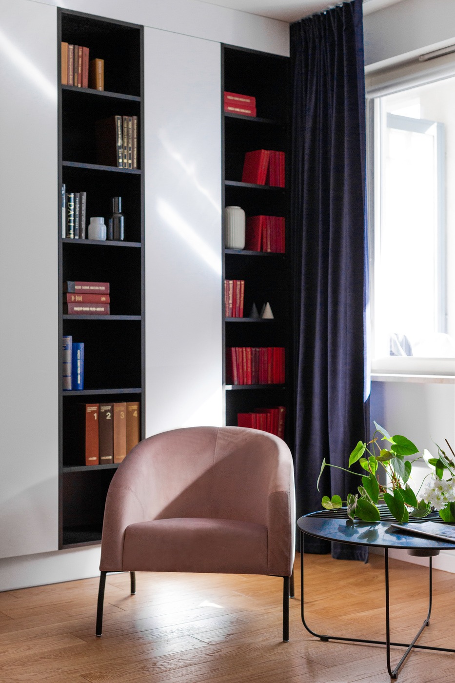 Jasnoróżowy fotel w minimalistycznym salonie z drewnianą podłogą, regałem z książkami i ciemnoniebieskimi zasłonami, doświetlonym promieniami słońca wpadającymi przez okno.
