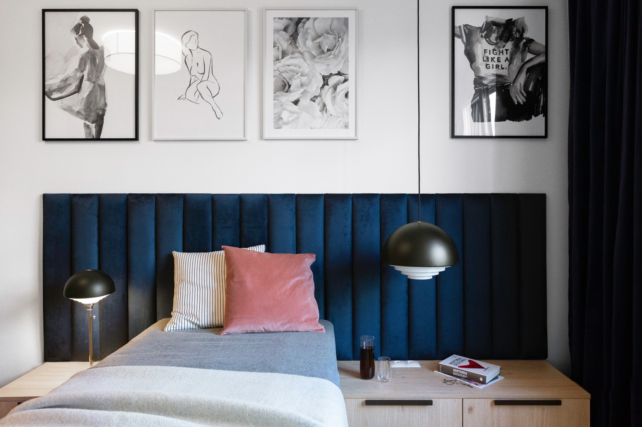 Sypialnia z ciemnoniebieskim, welurowym panelem zagłówkowym, minimalistycznymi grafikami na ścianie i drewnianą szafką nocną z książką 'Historia Designu'.