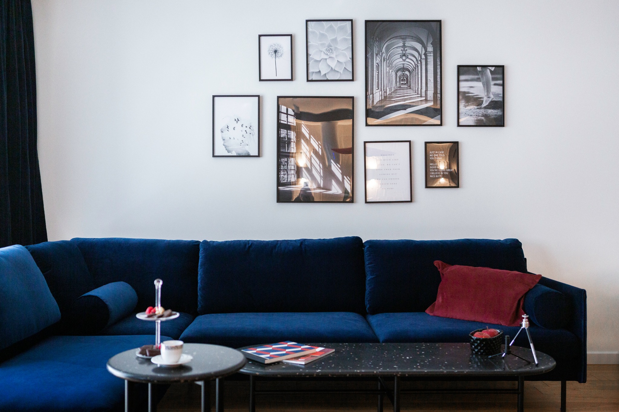 Salon w mieszkaniu na Żoliborzu, Warszawa. Granatowa sofa z poduszkami, stoliki kawowe z przekąskami i książkami, ściana ozdobiona galerią czarno-białych grafik w ramkach.