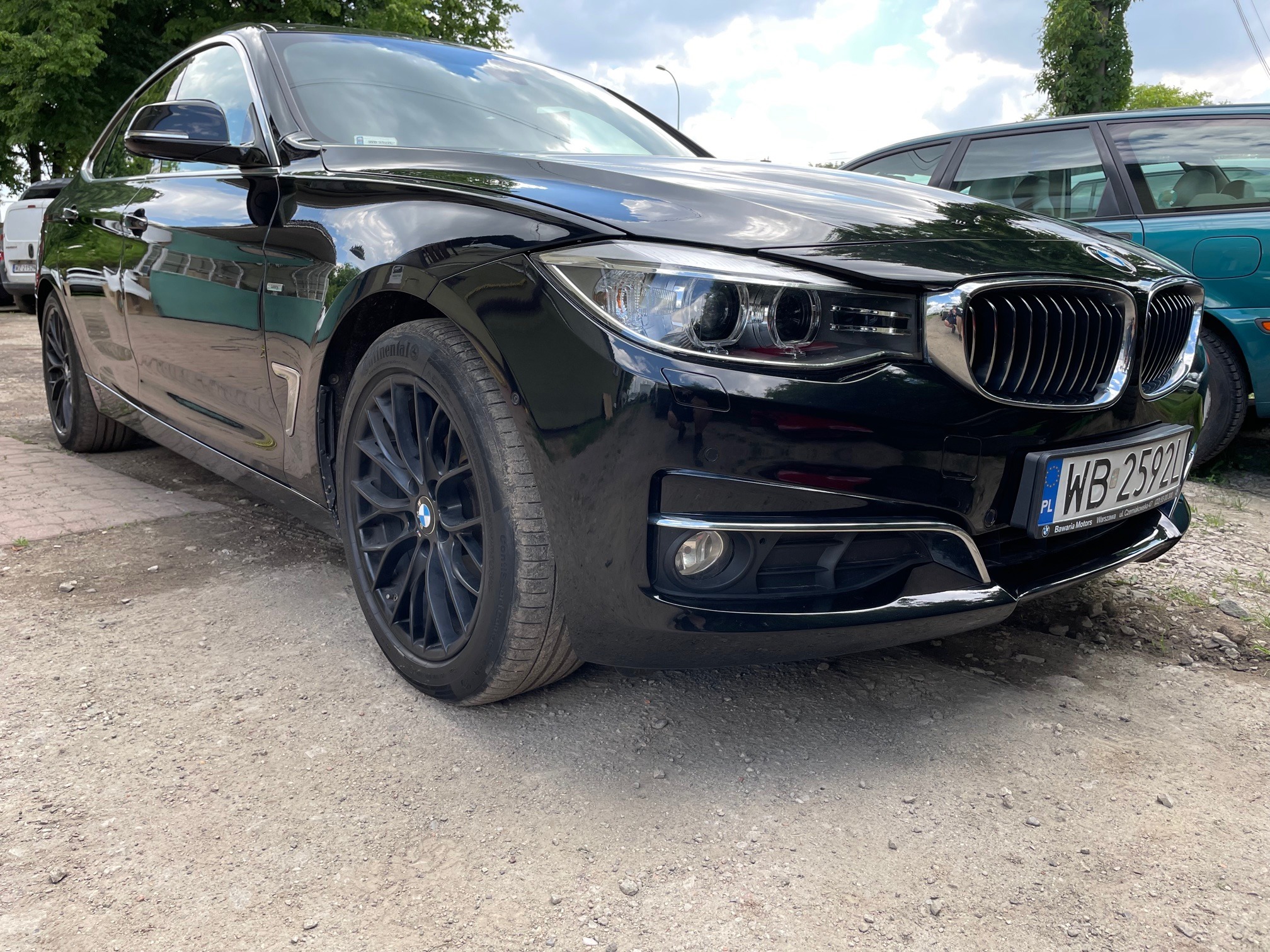 Czarny samochód BMW z aluminiowymi felgami, widoczny fragment karoserii z reflektorem i tablicą rejestracyjną na szutrowym podłożu.