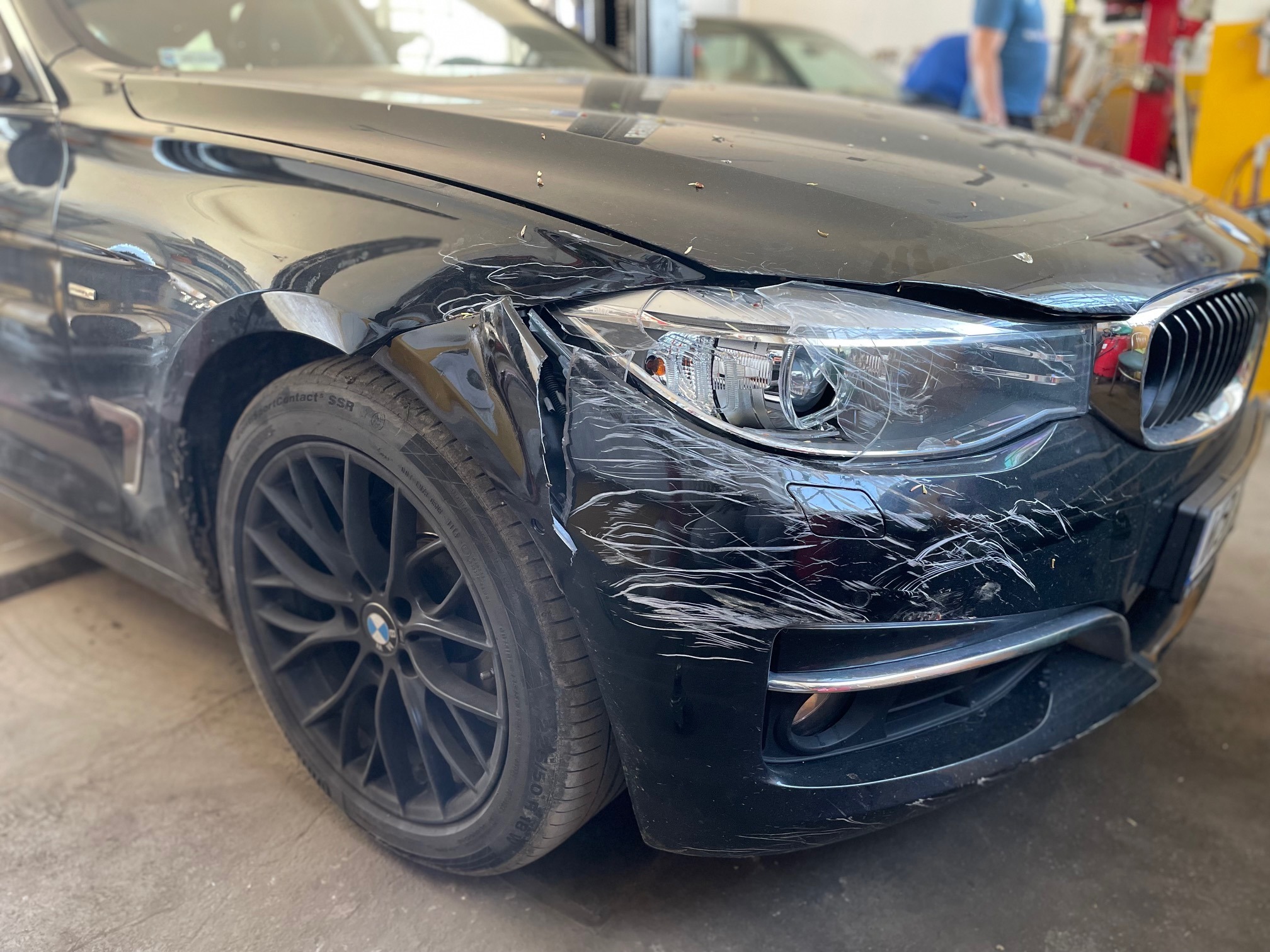 Uszkodzony przód czarnego samochodu marki BMW, widoczne wgniecenia i zarysowania na błotniku i reflektorze, zaparkowany w warsztacie.