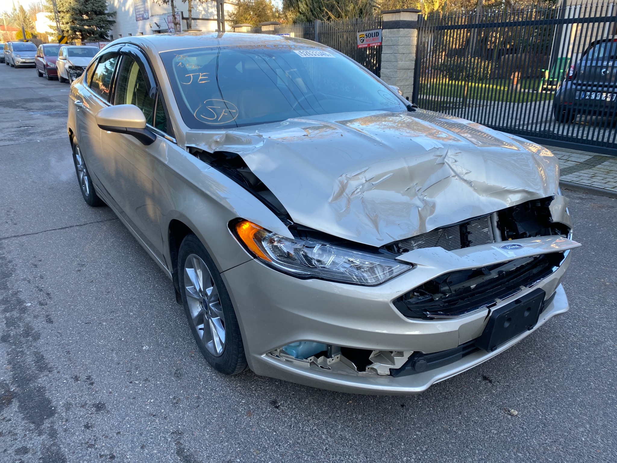 Uszkodzony przód złotego Forda Fusion z widocznymi zagnieceniami na masce, uszkodzonym zderzakiem i reflektorem, zaparkowany na ulicy z napisem 'JE SF' na przedniej szybie.