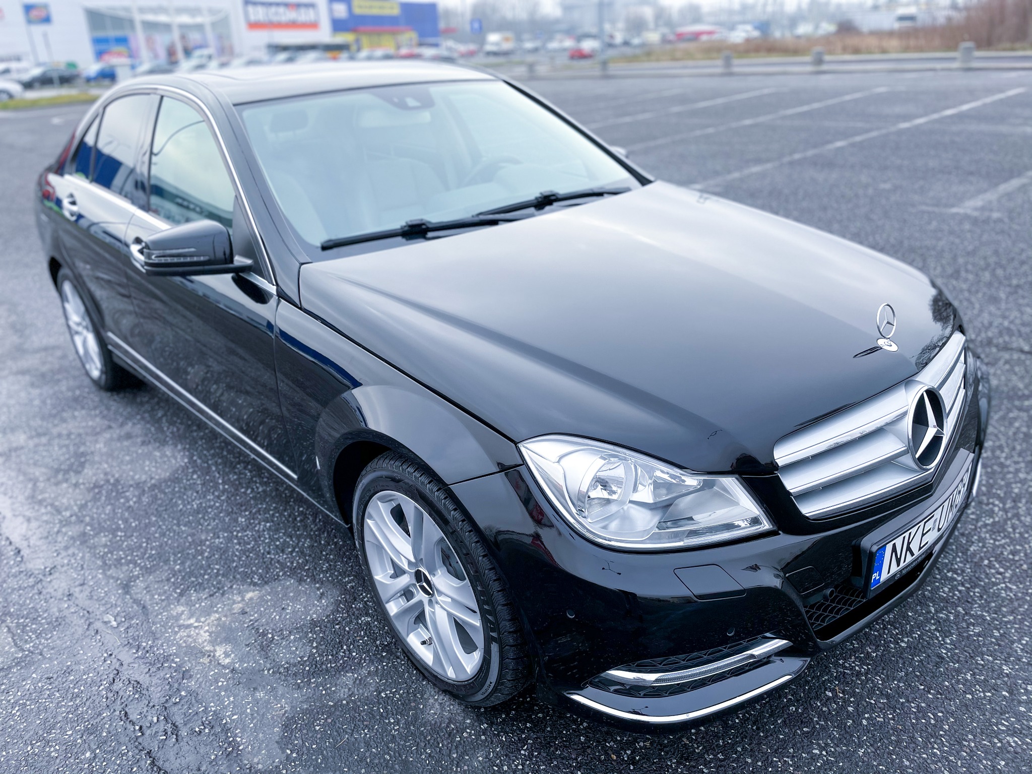Czarny samochód marki Mercedes-Benz klasy C, sfotografowany na parkingu przed centrum handlowym w pochmurny dzień. Widoczna polska tablica rejestracyjna.