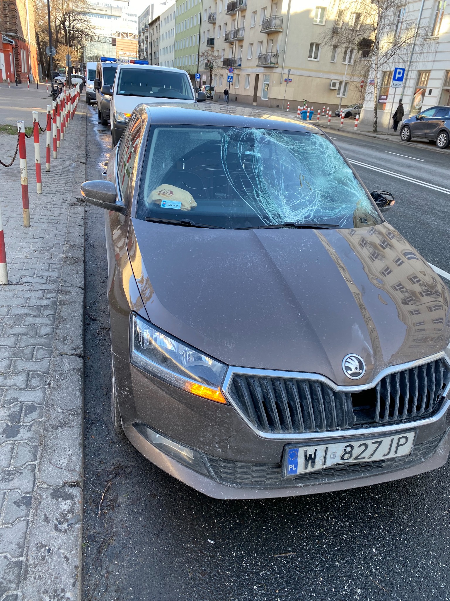 Uszkodzona przednia szyba brązowej Skody Fabii z widocznym pęknięciem na całej powierzchni, stojącej na ulicy z uszkodzonym zderzakiem i tablicą rejestracyjną WI 827JP.