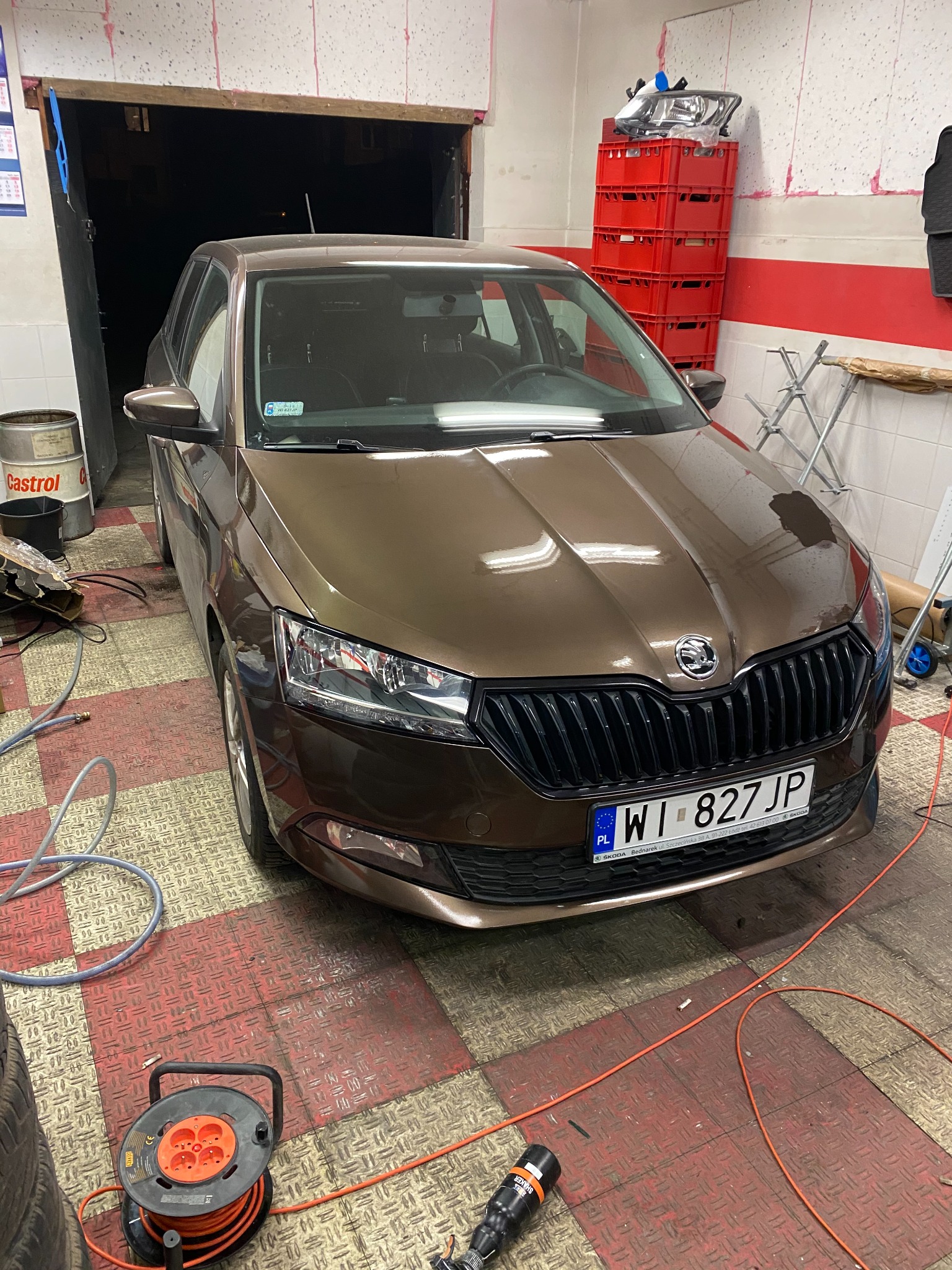 Brązowa Skoda Fabia po renowacji lakieru, zaparkowana w warsztacie z podłogą w kratkę, widoczny bęben z przedłużaczem i narzędzia.