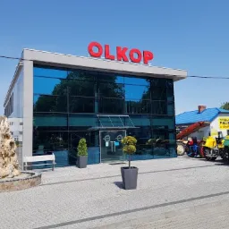 Budynek firmy OLKOP z czerwonym napisem na dachu, przeszklona fasada, przed wejściem fontanna z kamienia, obok zaparkowane maszyny budowlane i stacja paliw.