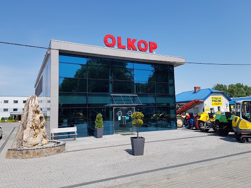Budynek firmy OLKOP z czerwonym napisem na dachu, przeszklona fasada, przed wejściem fontanna z kamienia, obok zaparkowane maszyny budowlane i stacja paliw.