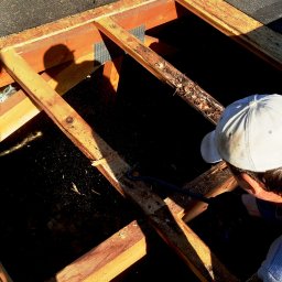 KMMJ Firma dekarska w Bielsku-Białej - Pracownik w czapce demontuje drewnianą konstrukcję dachu przy użyciu łomu, widok z góry na częściowo odsłonięty dach.