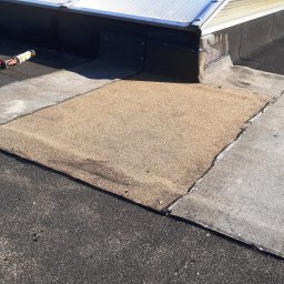 KMMJ Firma dekarska w Bielsku-Białej - Fragment płaskiego dachu z widocznymi warstwami papy, miejscem naprawy pokrytym drobnym kruszywem, narzędziami pracy: wiertarką, wiaderkiem i tubą z uszczelniaczem oraz świetlikiem dachowym w tle.