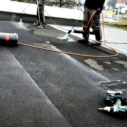 KMMJ Firma dekarska w Bielsku-Białej - Pracownicy podczas hydroizolacji dachu płaskiego za pomocą palnika dekarskiego, widoczny sprzęt i narzędzia pracy.