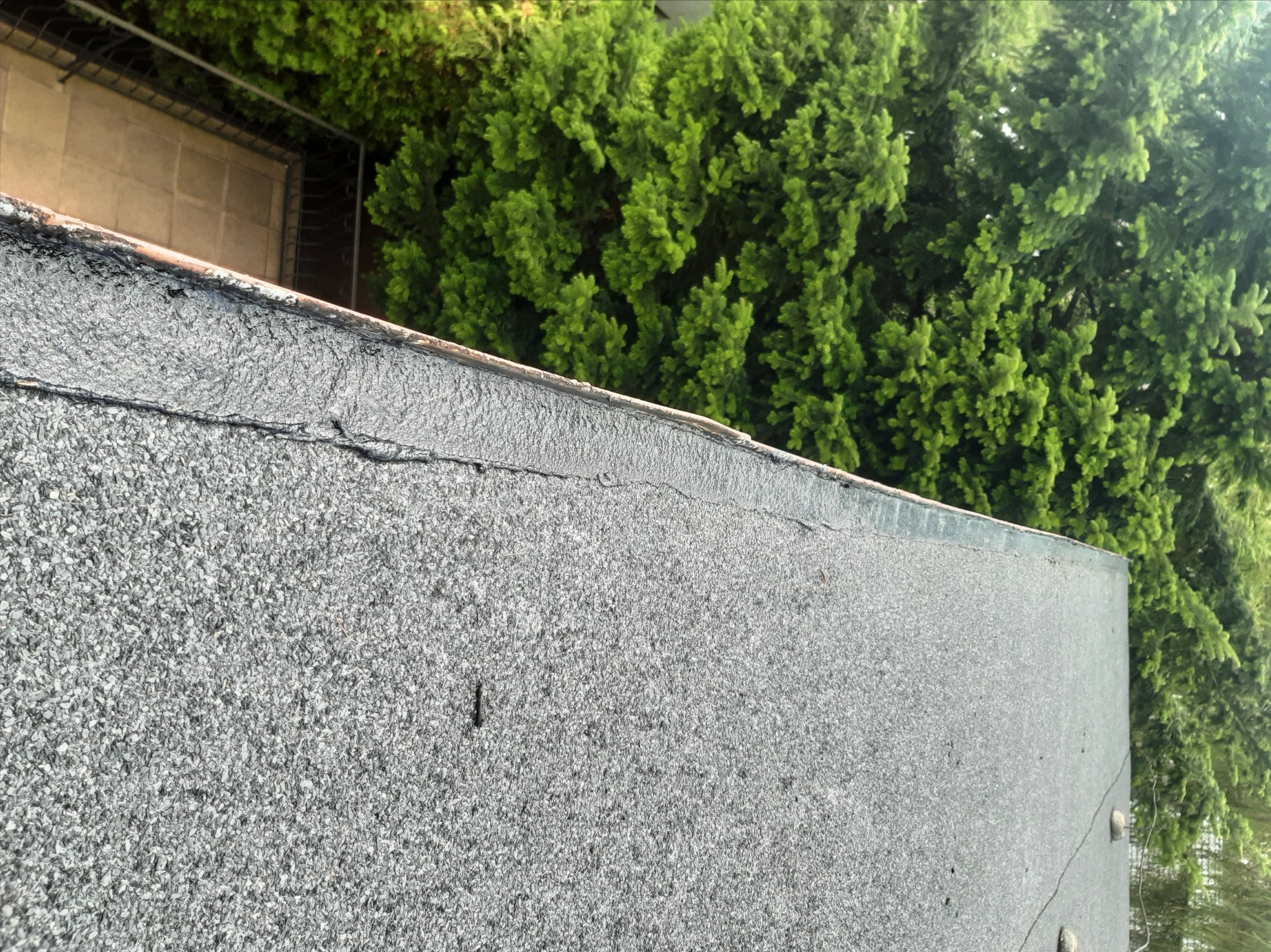 Szczegółowe ujęcie płaskiego dachu pokrytego papą z posypką mineralną, z widocznym połączeniem z obróbką blacharską i bujną zielenią drzew w tle.