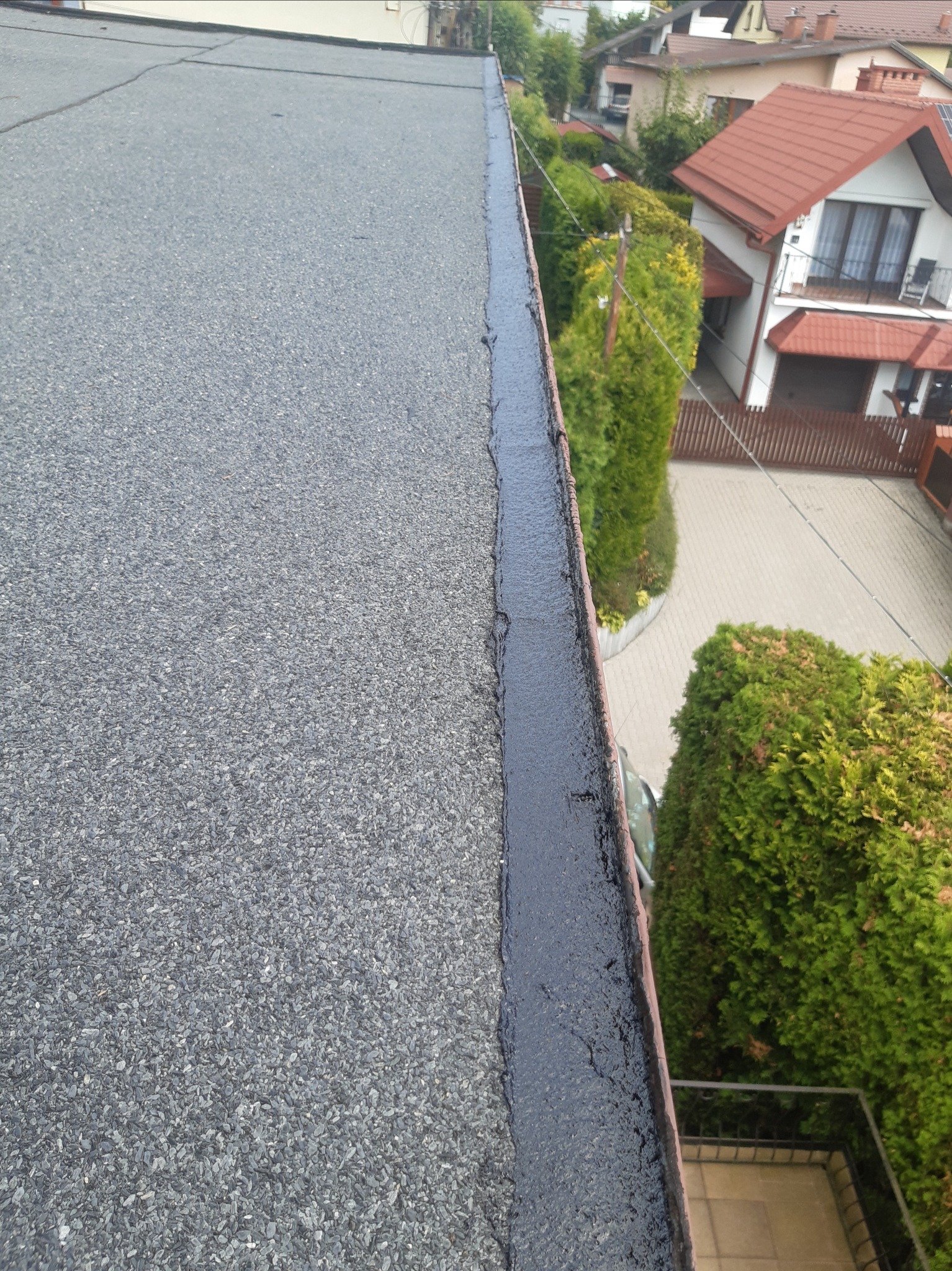 Widok z góry na płaski dach pokryty papą, z widocznymi obróbkami blacharskimi i uszczelnieniami, w tle zabudowa jednorodzinna i zieleń.