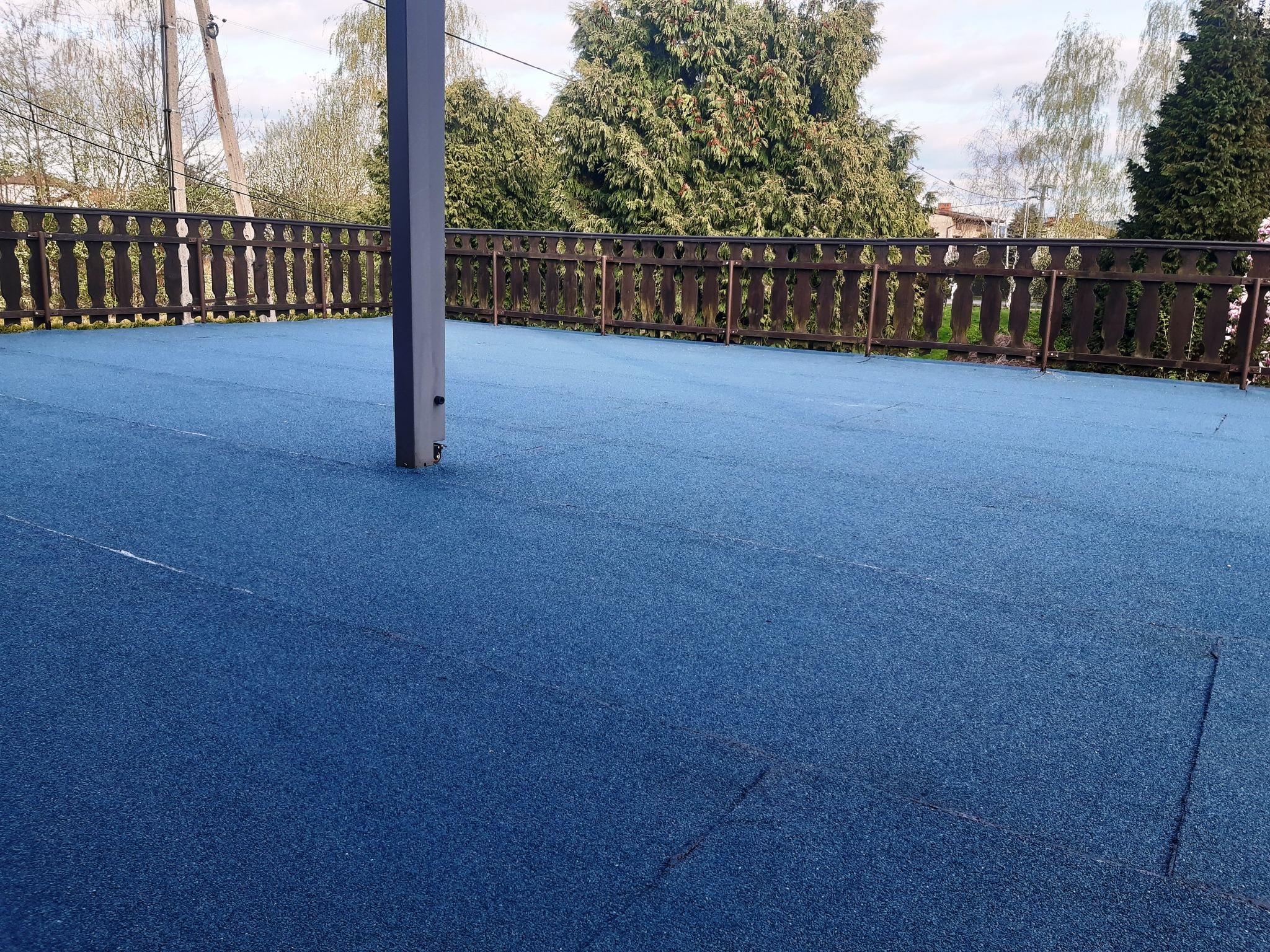 Niebieska, płaska powierzchnia dachu tarasu z drewnianą balustradą i metalowym słupem, widok na zieleń drzew w tle.