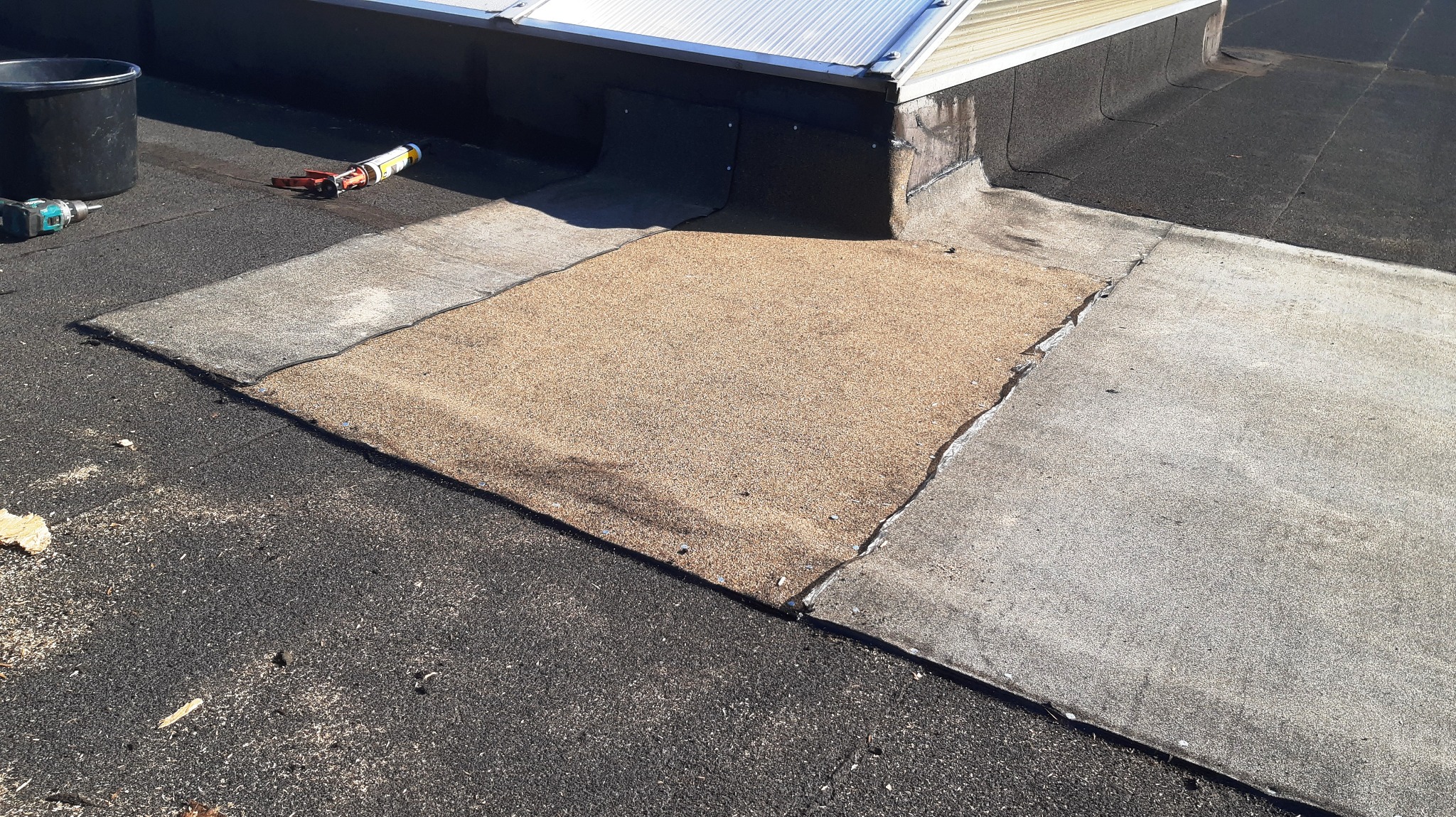 Fragment płaskiego dachu z widocznymi warstwami papy, miejscem naprawy pokrytym drobnym kruszywem, narzędziami pracy: wiertarką, wiaderkiem i tubą z uszczelniaczem oraz świetlikiem dachowym w tle.