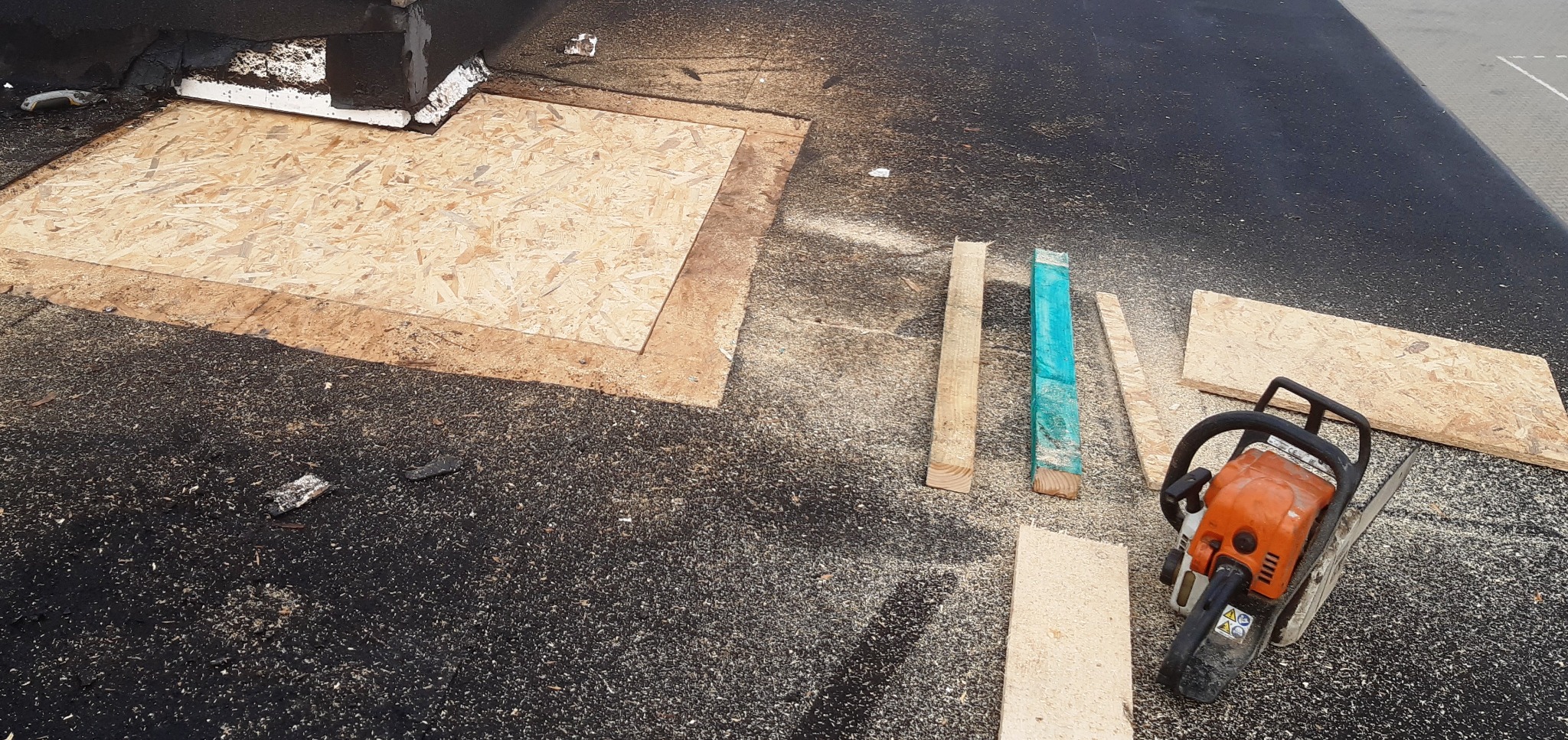 Fragment dachu z deską OSB, świeżymi wiórami i piłą łańcuchową, widoczne także drewniane łatki, jedna pomalowana na turkusowo.