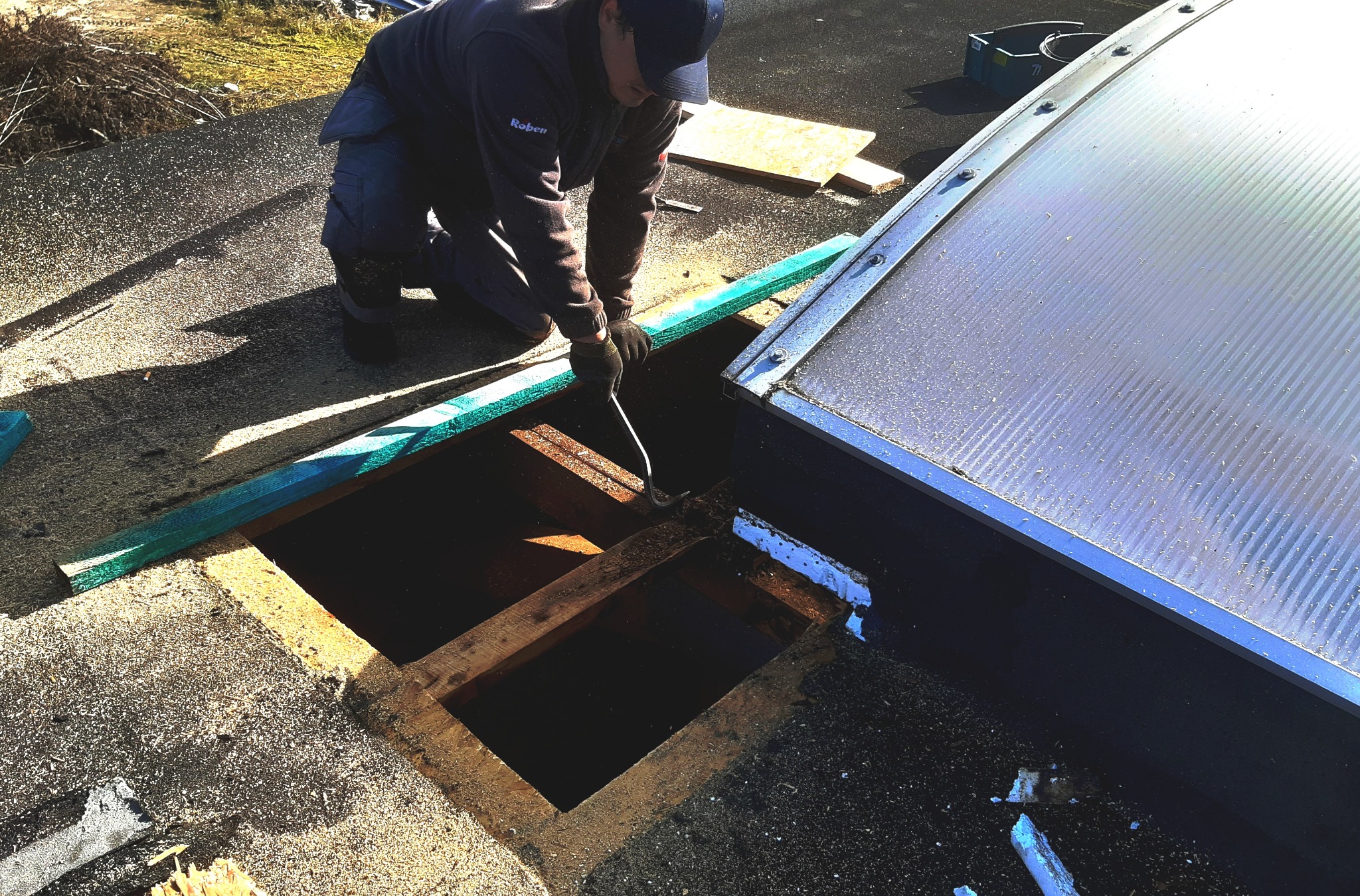 Dekarz w trakcie naprawy dachu z użyciem łomu, widoczny otwór w poszyciu dachu i fragment świetlika dachowego