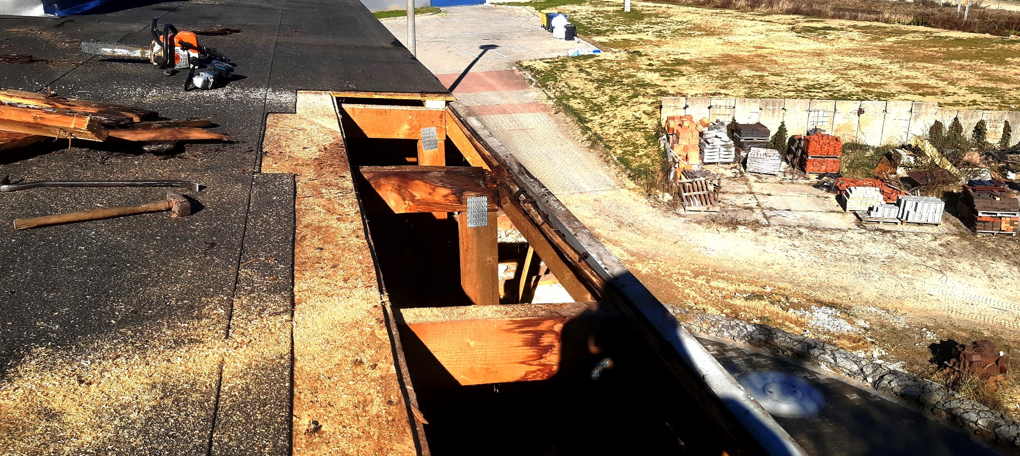 Demontaż starego dachu z widoczną konstrukcją więźby dachowej, leżące narzędzia: piła spalinowa, młotek i łom, w tle plac z materiałami budowlanymi.