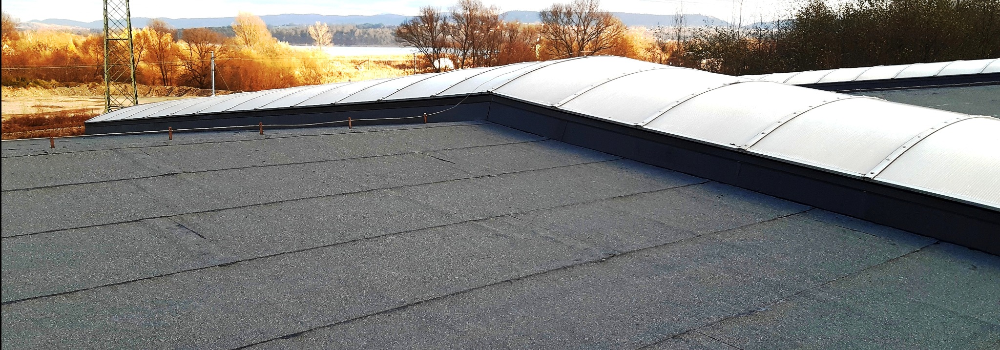 Widok z góry na płaski dach pokryty papą, z zamontowanymi świetlikami tunelowymi i wystającymi elementami odgromowymi, w tle widoczne drzewa i krajobraz.