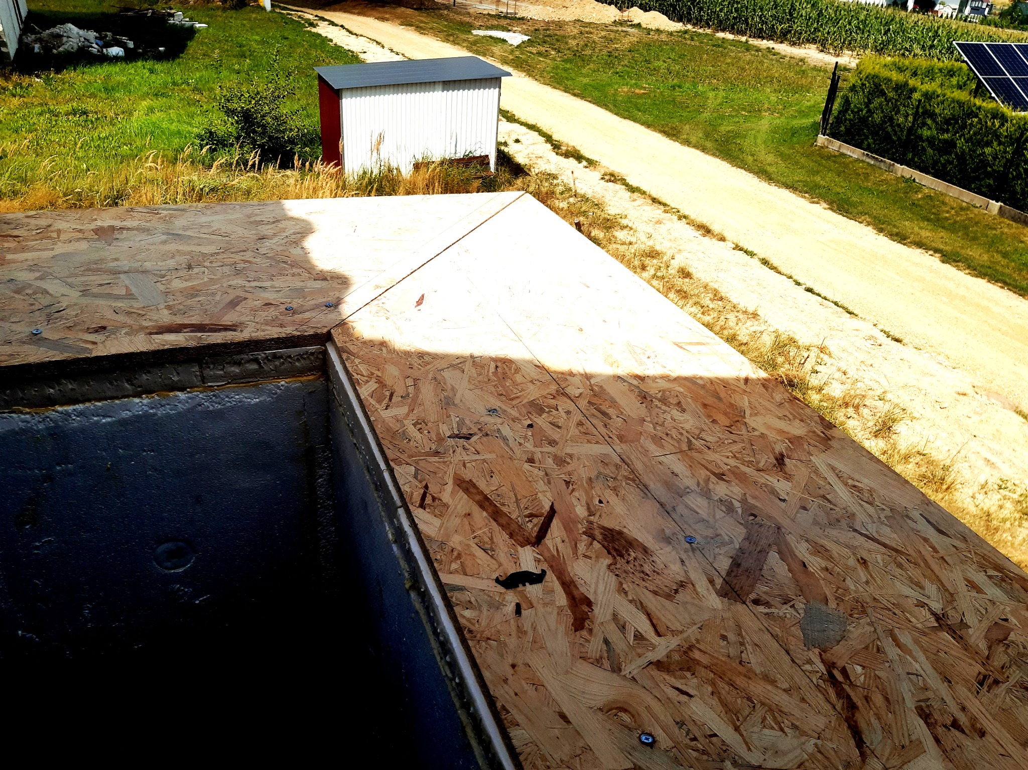 Widok z góry na fragment dachu w trakcie budowy, z widoczną płytą OSB przymocowaną do konstrukcji, w tle budynek gospodarczy, droga gruntowa i pole uprawne.