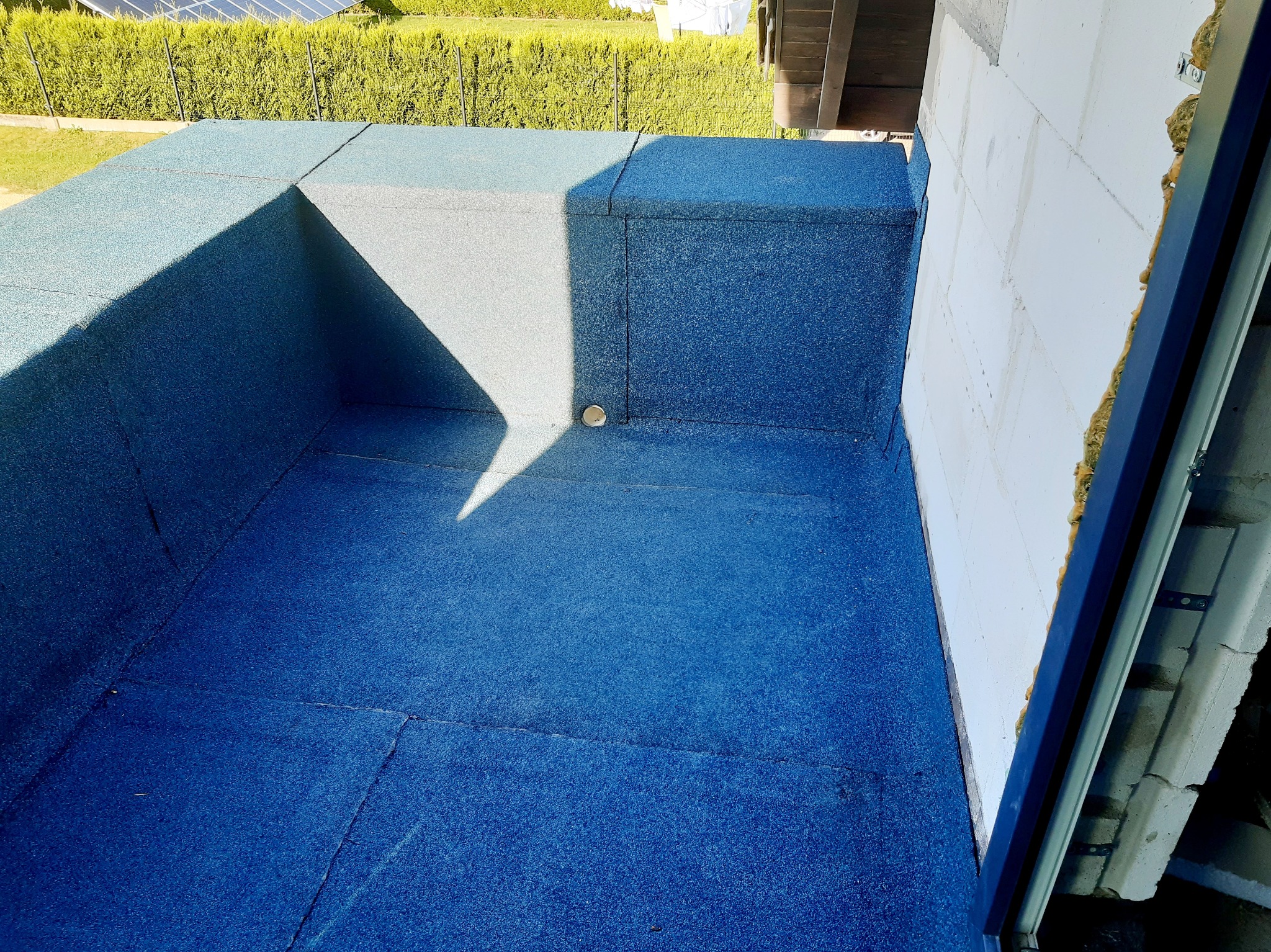 Płaski dach pokryty niebieską papą termozgrzewalną, widoczny kominek wentylacyjny i fragment ściany budynku z ociepleniem.