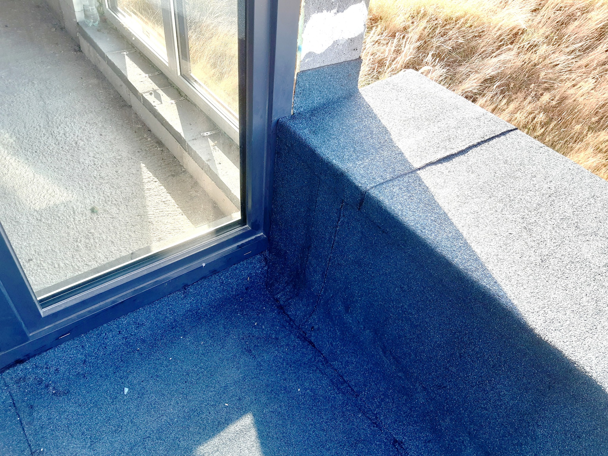 Szczegółowe ujęcie hydroizolacji bitumicznej na płaskim dachu przy oknie, widoczne połączenie z ramą okienną i fragment otaczającego krajobrazu z trawami.