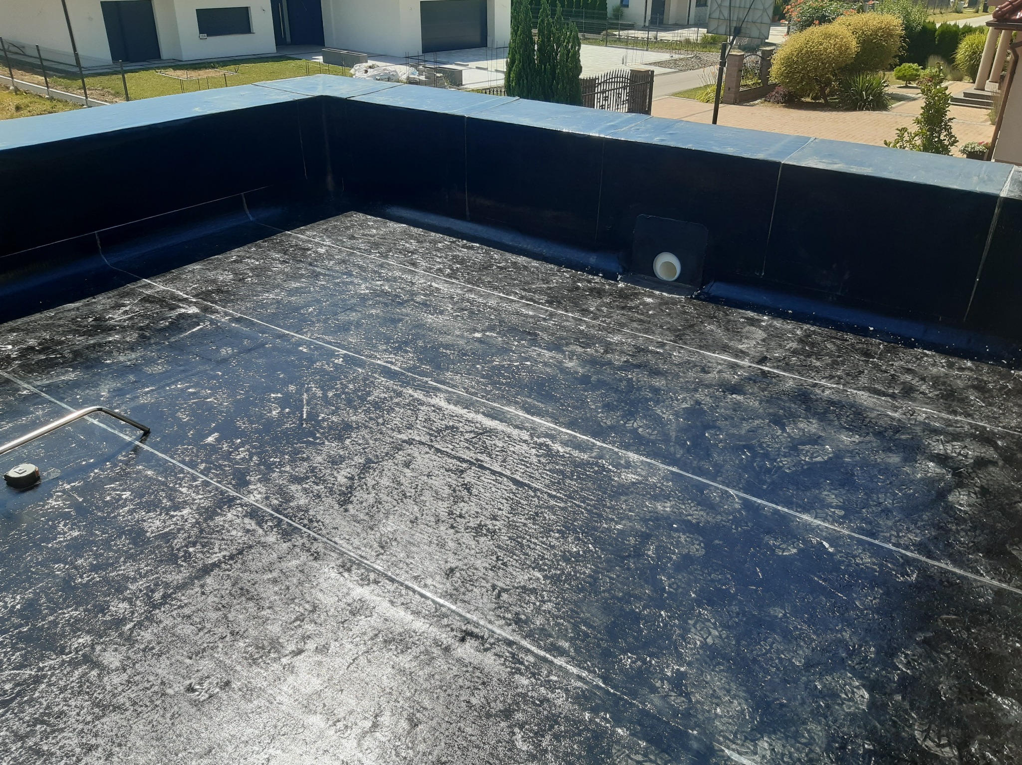 Płaski dach pokryty czarną membraną bitumiczną z widocznym odpływem wody deszczowej i narzędziem dekarskim leżącym na powierzchni, w tle budynki mieszkalne.