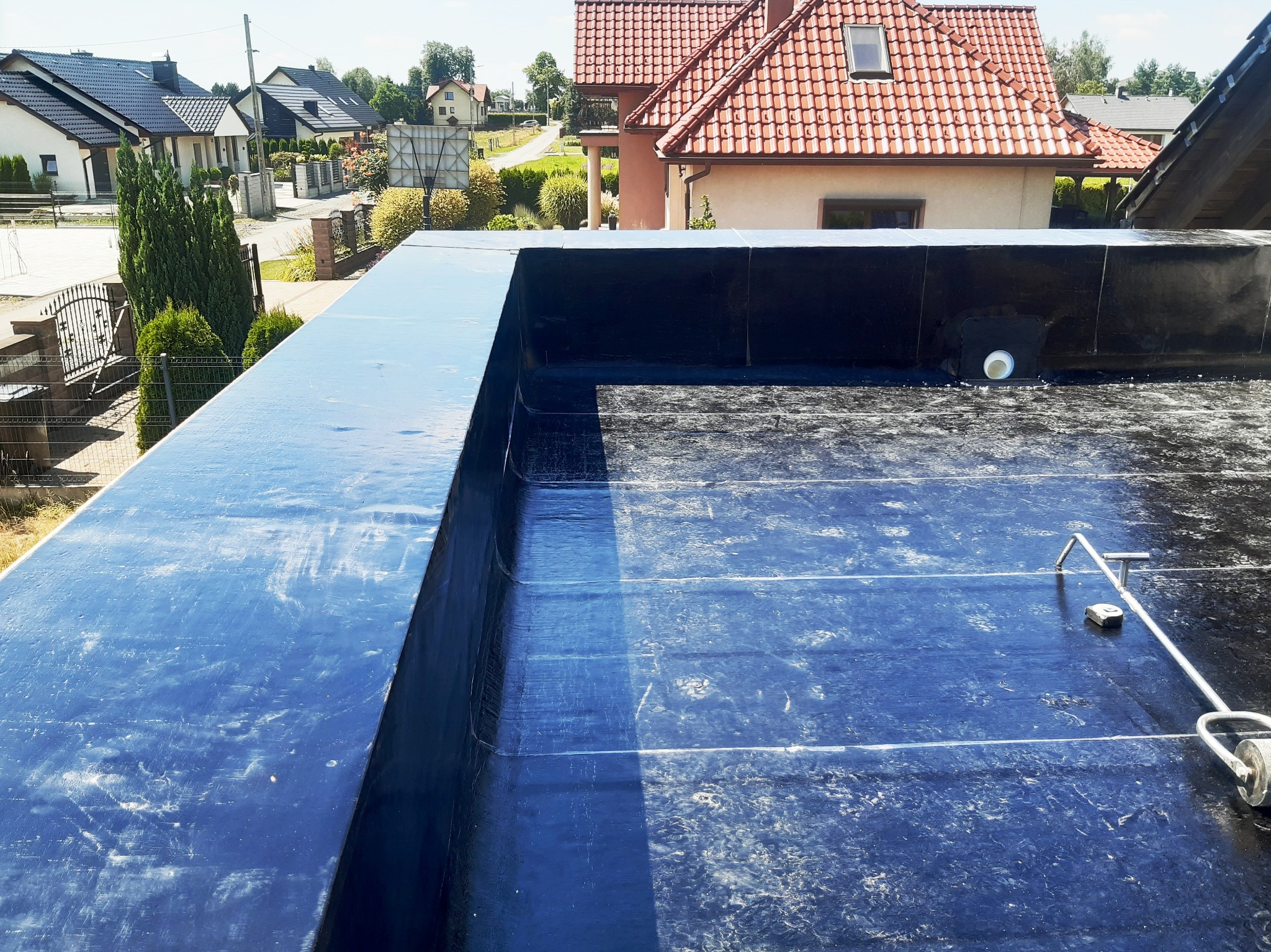 Widok z góry na płaski dach pokryty czarną membraną bitumiczną, z widocznym kominkiem wentylacyjnym i narzędziem do zgrzewania papy na tle zabudowy jednorodzinnej.