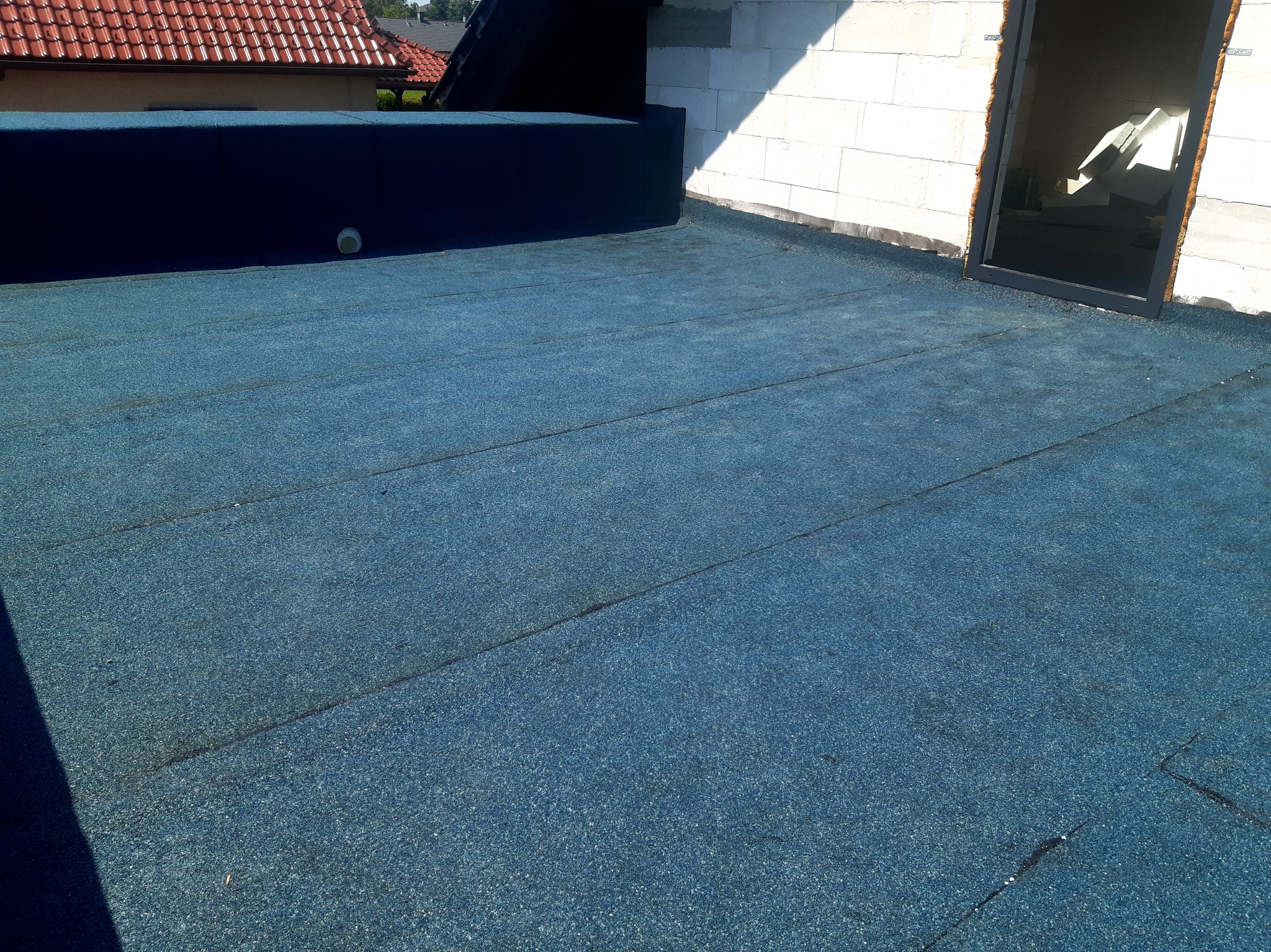 Nowe, niebieskie pokrycie dachu płaskiego z widoczną w oddali konstrukcją budynku i oknem dachowym w trakcie montażu.