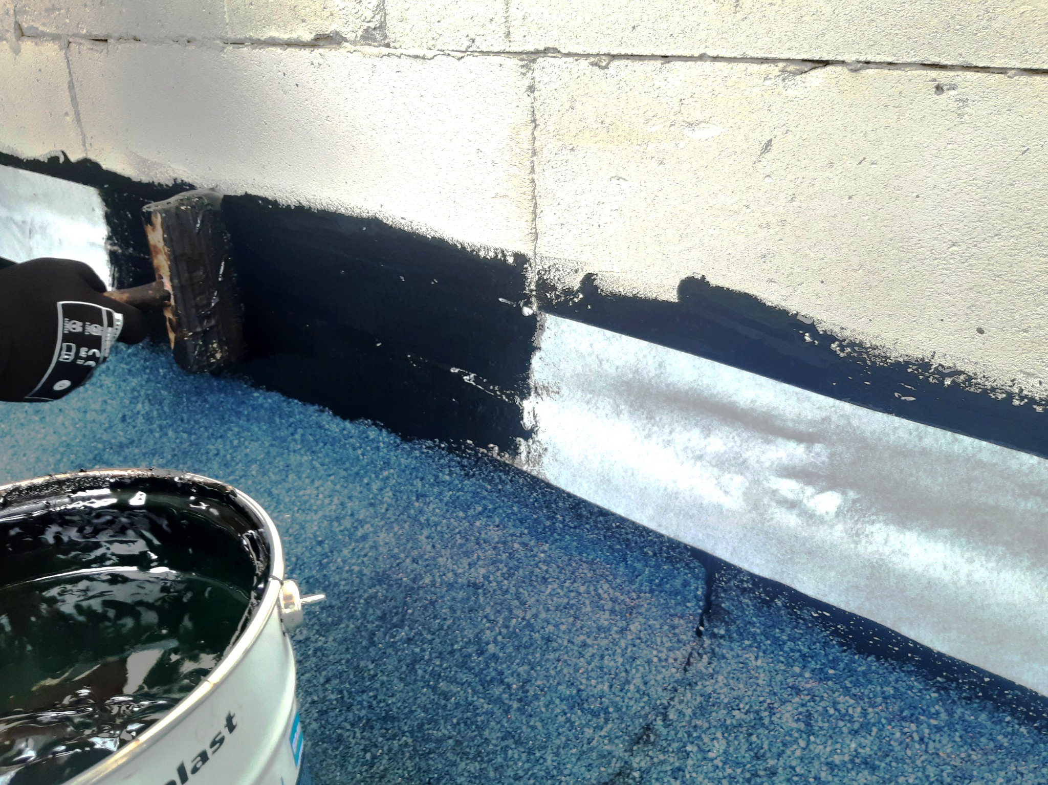 Hydroizolacja fundamentu z użyciem pędzla i czarnej substancji bitumicznej, widoczny fragment ściany fundamentowej z betonu komórkowego i wiadro z preparatem.