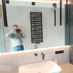 Nowoczesna łazienka z podświetlanym lustrem, odbijającym osobę robiącą zdjęcie, widoczny grzejnik drabinkowy, umywalka nablatowa i grafitowe płytki.