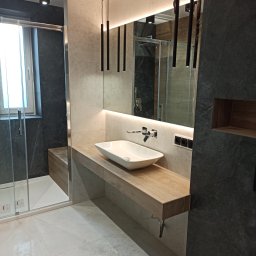 Delta - Nowoczesna łazienka z kabiną prysznicową, umywalką nablatową na drewnianym blacie, podświetlanym lustrem i wiszącymi lampami tubami.