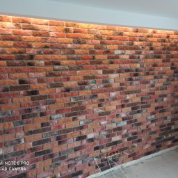 Delta - Ściana wyłożona dekoracyjną cegłą w odcieniach czerwieni i brązu, z podświetleniem LED u góry, widoczne przewody elektryczne w dolnej części ściany i skrzynka narzędziowa w rogu pomieszczenia.