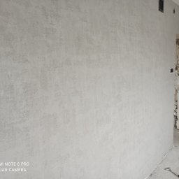 Delta - Ściana pokryta szarą, dekoracyjną strukturą tynku w nowym mieszkaniu, widoczny fragment ściany z cegły w przejściu, surowy beton na podłodze.