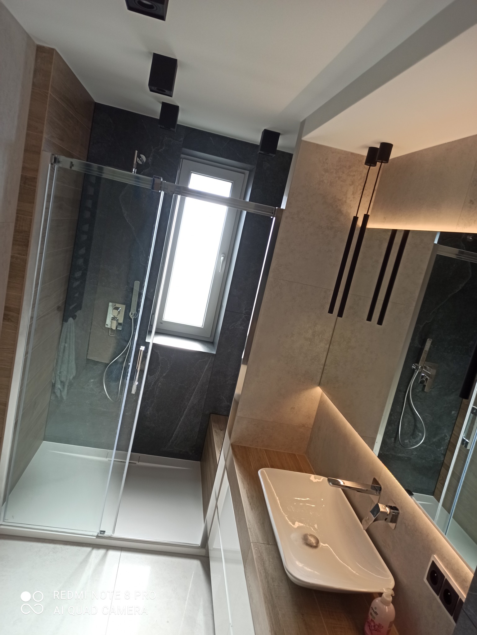 Nowoczesna łazienka z kabiną prysznicową, umywalką nablatową i oświetleniem LED pod lustrem. Czarne płytki imitujące kamień na ścianie prysznica, kontrastujące z jasnymi płytkami na pozostałej...