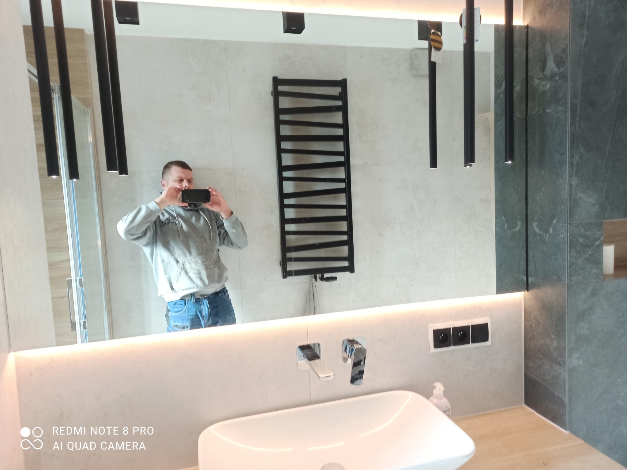 Nowoczesna łazienka z podświetlanym lustrem, odbijającym osobę robiącą zdjęcie, widoczny grzejnik drabinkowy, umywalka nablatowa i grafitowe płytki.