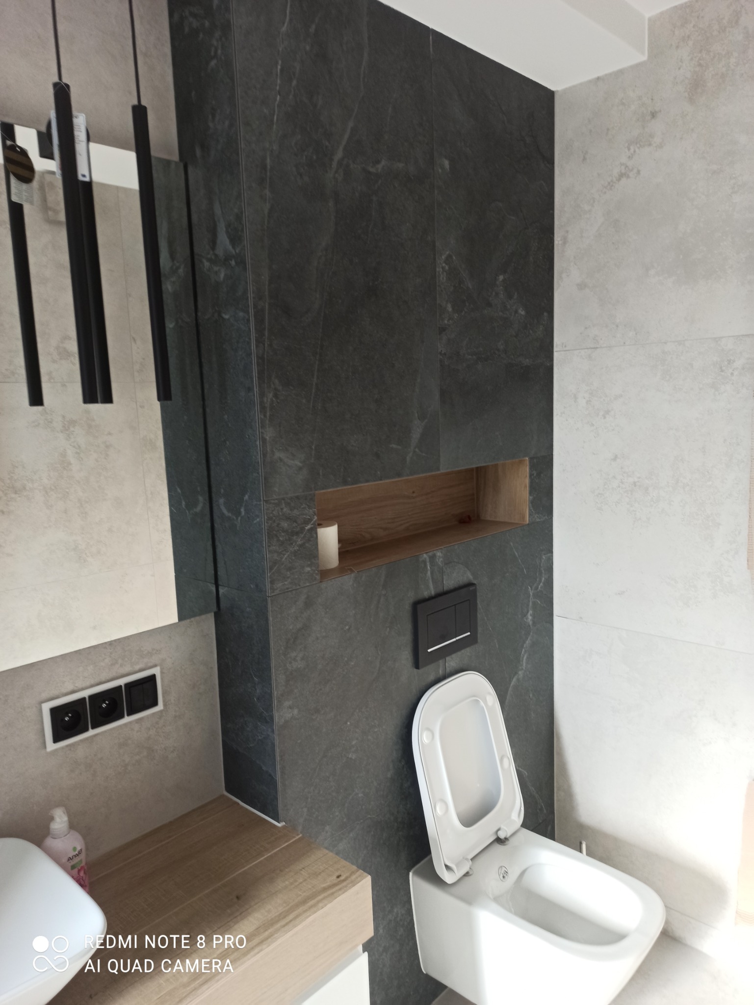 Nowoczesna łazienka z ciemnymi płytkami imitującymi kamień, drewnianą półką w zabudowie i podwieszaną toaletą z otwartą klapą.