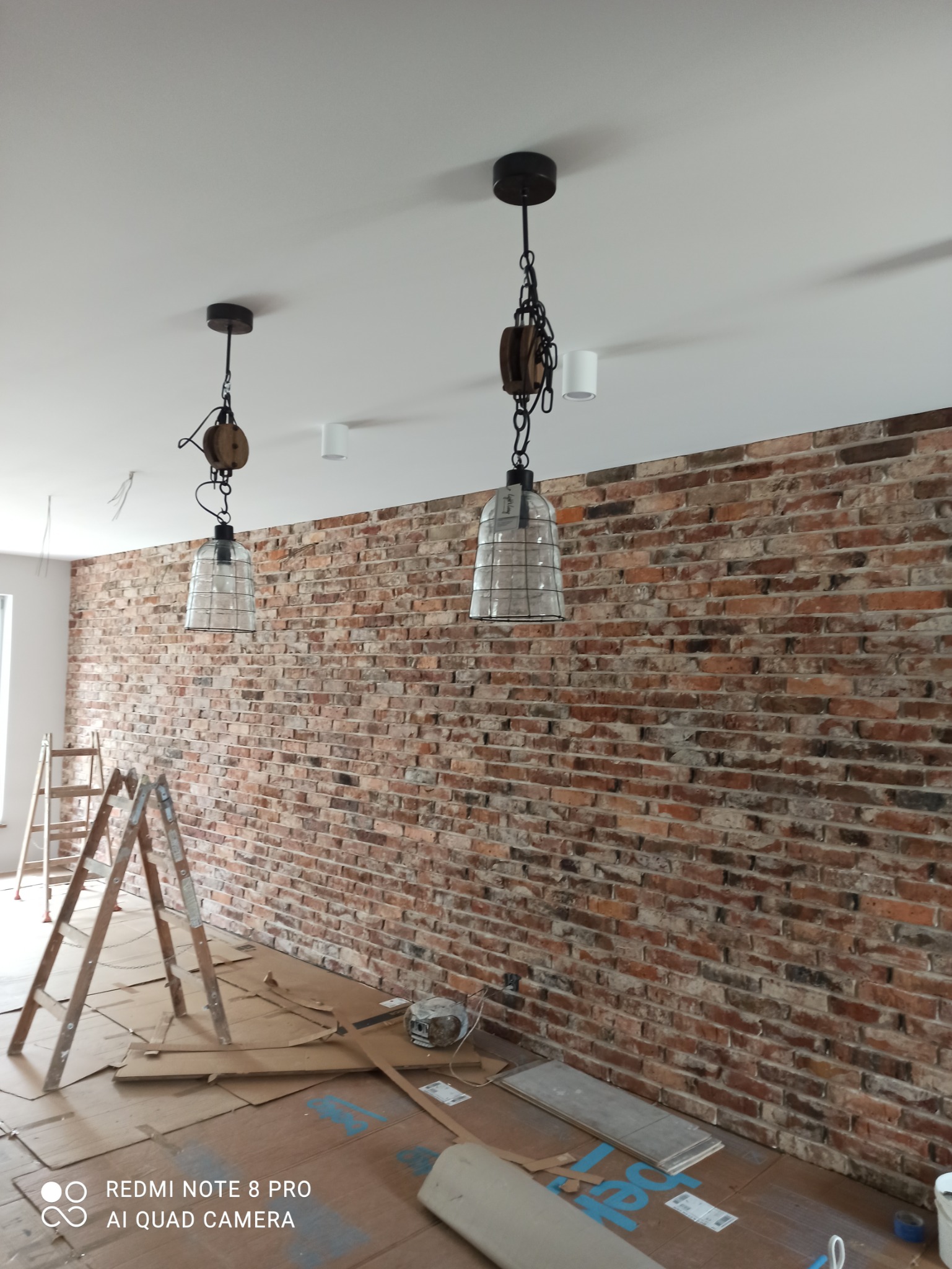 Ściana z czerwonej cegły z dekoracyjnymi lampami wiszącymi na tle zabezpieczonej podłogi i drabiny, sugerujące prace remontowe w trakcie.