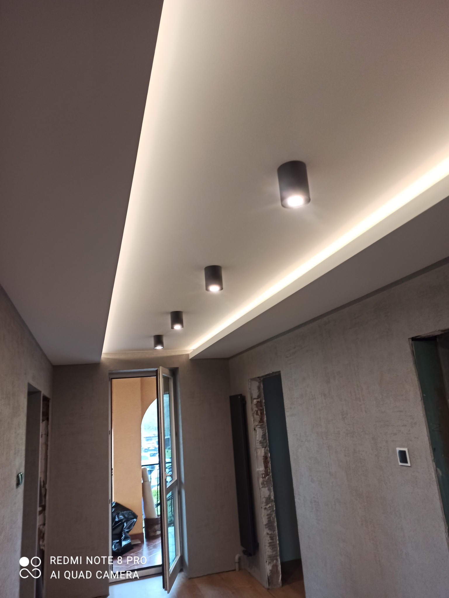 Wykończone wnętrze z podwieszonym sufitem z oświetleniem LED i punktowymi lampami, widoczne surowe ściany i otwarte przejścia drzwiowe.