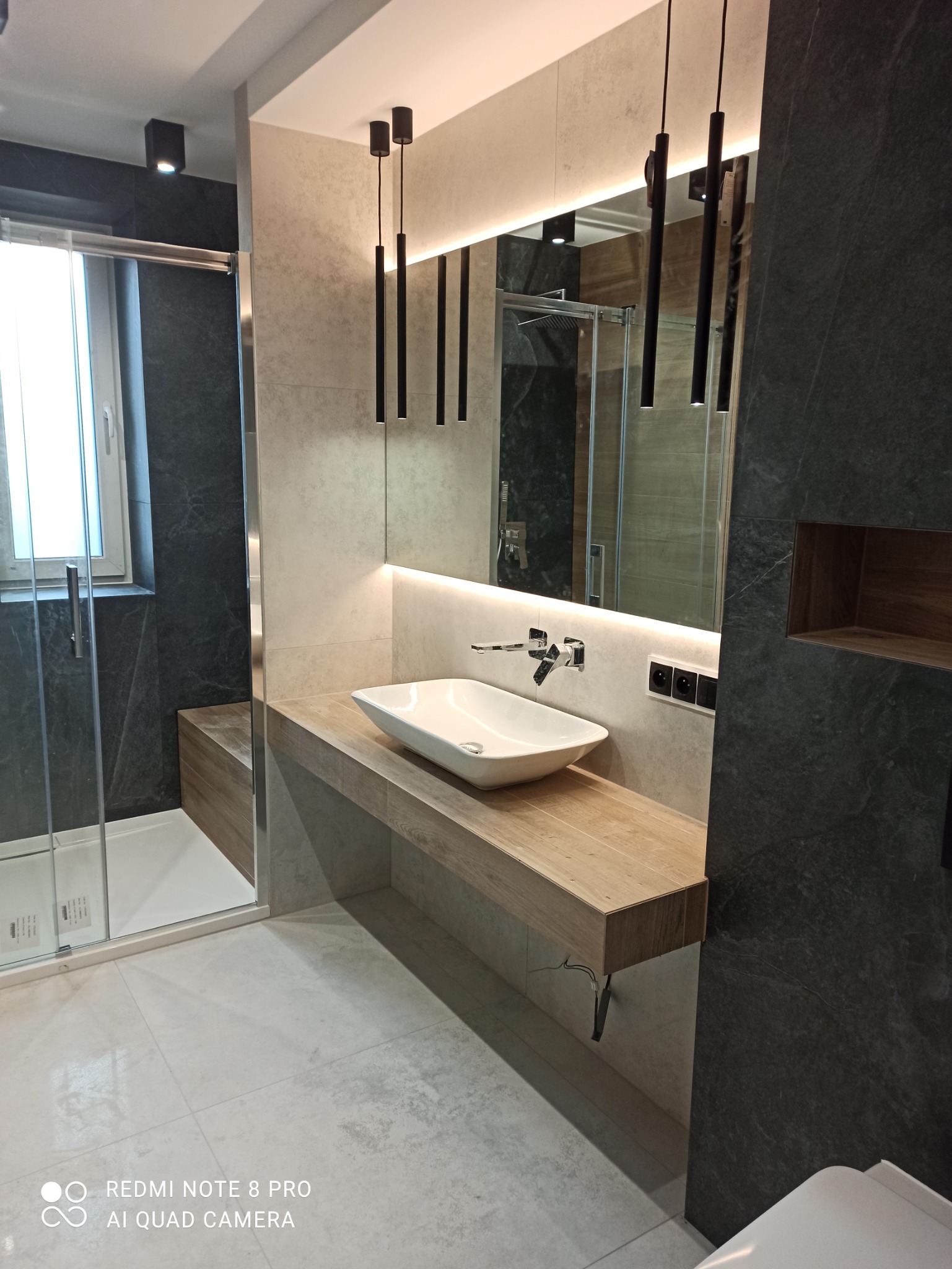 Nowoczesna łazienka z kabiną prysznicową, umywalką nablatową na drewnianym blacie, podświetlanym lustrem i wiszącymi lampami tubami.