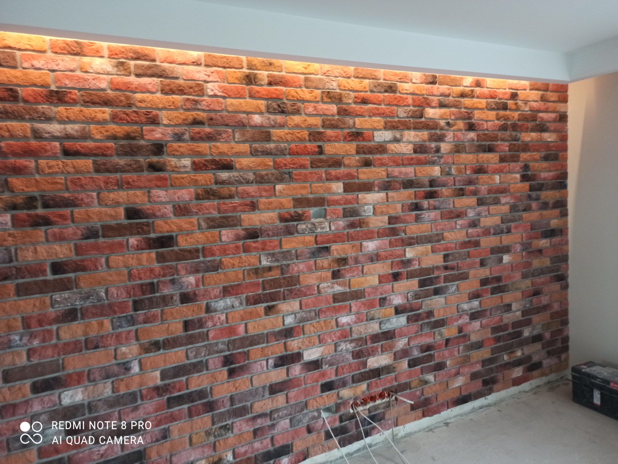 Ściana wyłożona dekoracyjną cegłą w odcieniach czerwieni i brązu, z podświetleniem LED u góry, widoczne przewody elektryczne w dolnej części ściany i skrzynka narzędziowa w rogu pomieszczenia.