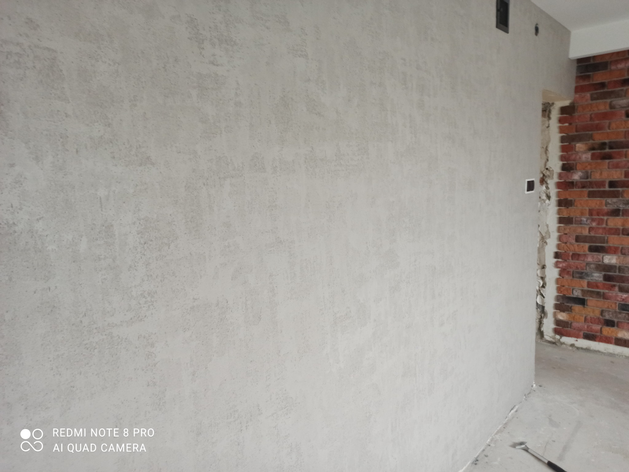 Ściana pokryta szarą, dekoracyjną strukturą tynku w nowym mieszkaniu, widoczny fragment ściany z cegły w przejściu, surowy beton na podłodze.