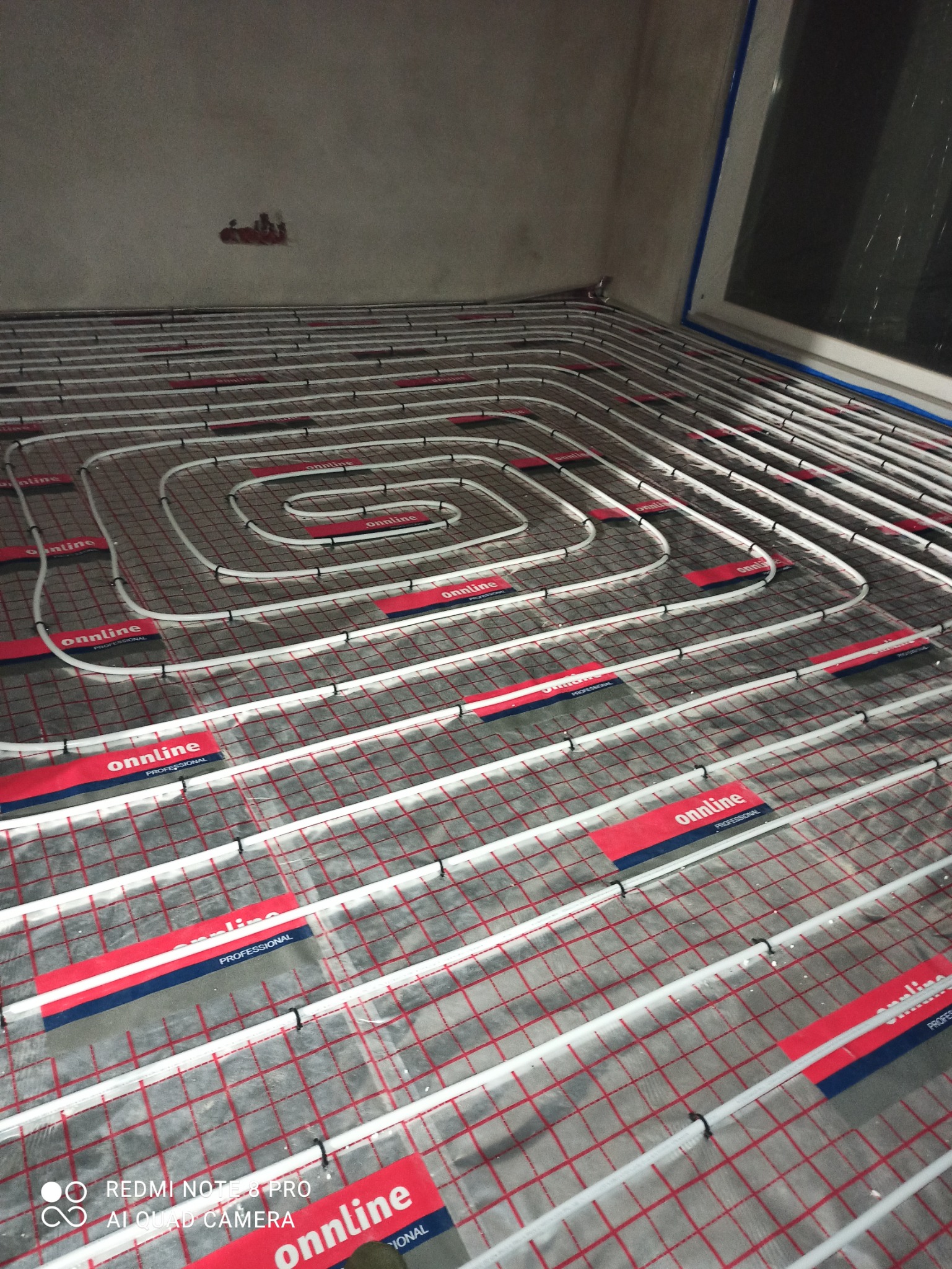 Instalacja ogrzewania podłogowego z rur rozprowadzonych na siatce, widoczne mocowania i etykiety producenta na folii, surowy stan ścian i okno w tle.