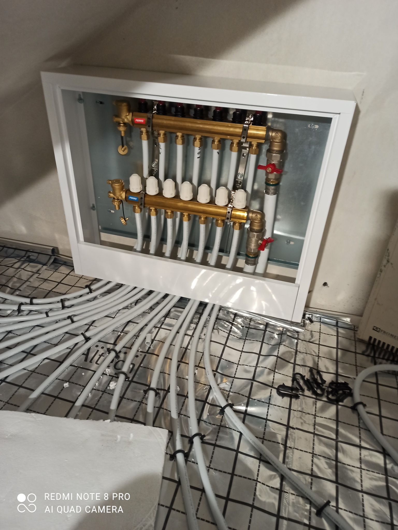 Rozdzielacz ogrzewania podłogowego Ferro w białej obudowie, białe rury grzewcze ułożone na folii z rastrem, czarne spinki mocujące rury.