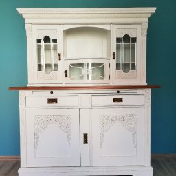 Pracownia Tapicerska Meblo-Komfort - Biały, dwuczęściowy kredens z drewnianym blatem, zdobiony rzeźbieniami, z przeszklonymi górnymi szafkami i szufladami, na tle turkusowej ściany i drewnianej podłogi.