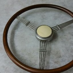 Pracownia Tapicerska Meblo-Komfort - Klasyczna kierownica samochodowa z drewnianym wieńcem, metalowymi szprychami i kremowym środkiem, leżąca na szarym tle.