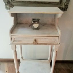 Pracownia Tapicerska Meblo-Komfort - Biała, odrestaurowana szafka nocna w stylu vintage z szufladą i półką, na której stoi szklany słoik z ozdobnym sercem, w tle fragment ramy obrazu.