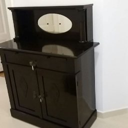 Pracownia Tapicerska Meblo-Komfort - Ciemnobrązowa, lakierowana komoda z dwoma drzwiczkami, szufladą i nadstawką z owalnym lustrem, widoczny klucz w zamku szuflady, ustawiona przy białej ścianie.