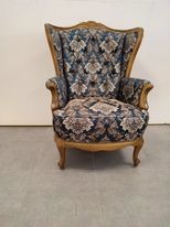 Odnowiony fotel z wysokim oparciem, w niebiesko-brązowe wzory kwiatowe, z drewnianą, rzeźbioną ramą w kolorze jasnego drewna, stojący na szarej podłodze na tle białej ściany.