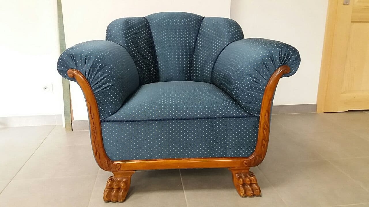 Stylowy, odnowiony fotel w niebieskiej tapicerce w białe kropki, z drewnianymi, rzeźbionymi podłokietnikami i nóżkami w kształcie lwich łap, na tle jasnych płytek podłogowych i drzwi.
